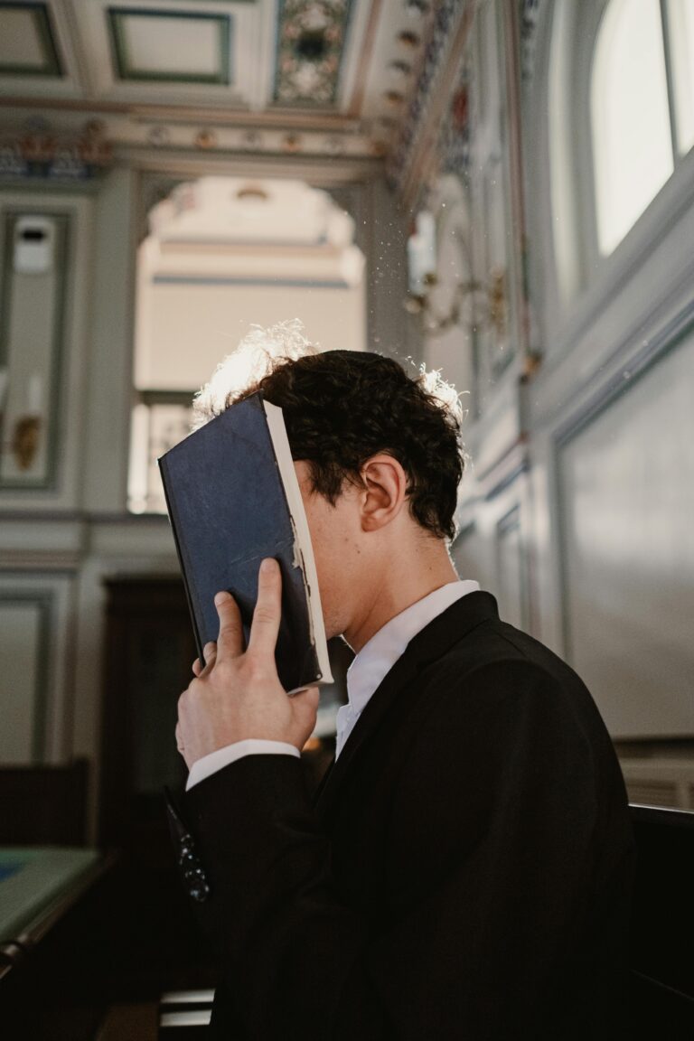 A young man in a suit prays with a book in a sunlit synagogue, capturing a moment of faith.